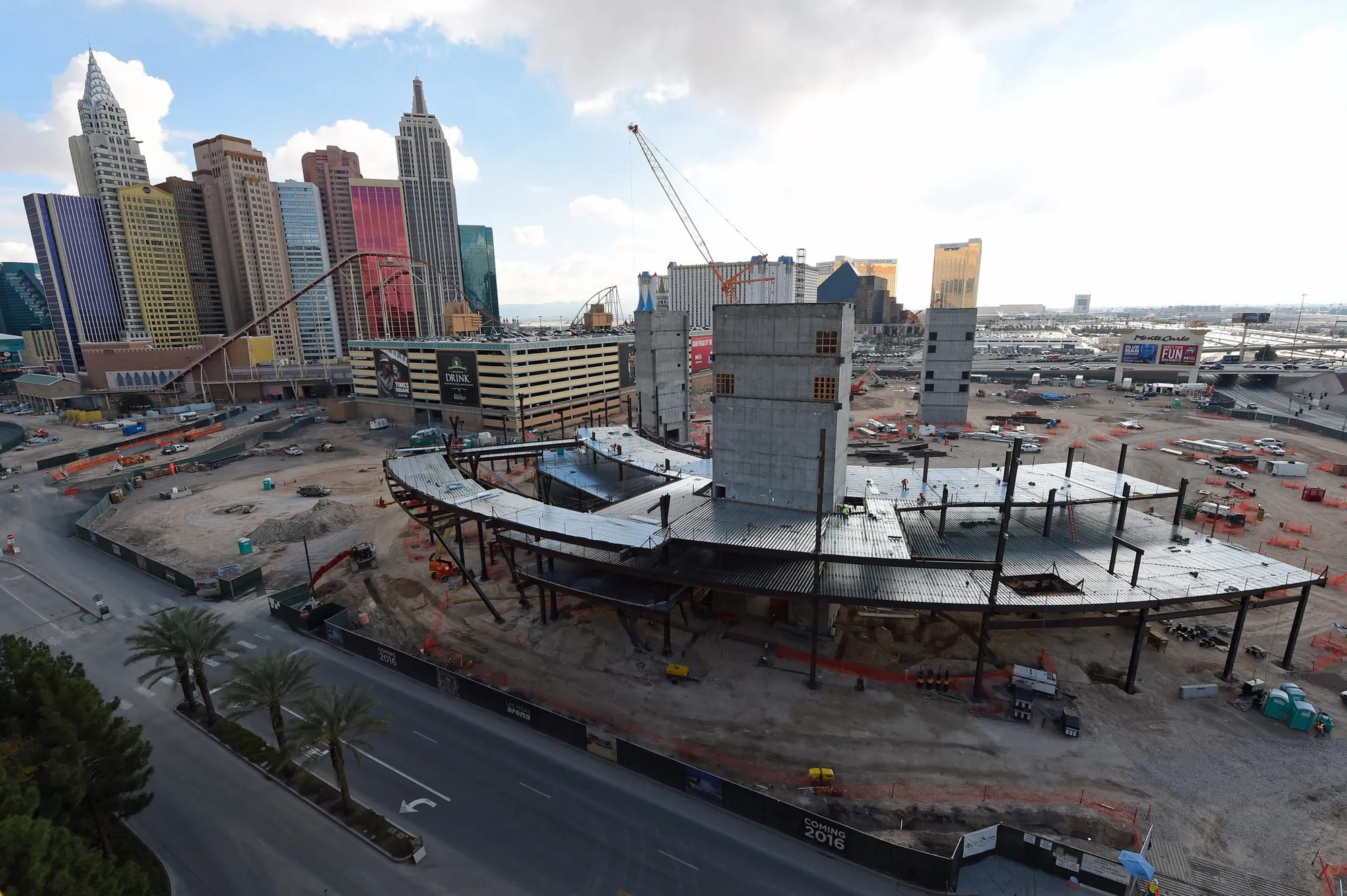T-Mobile Arena under construction, steel frame and concrete cores rising on a bare site with the Las Vegas Strip skyline behind it and a barricade wrap reading COMING 2016.