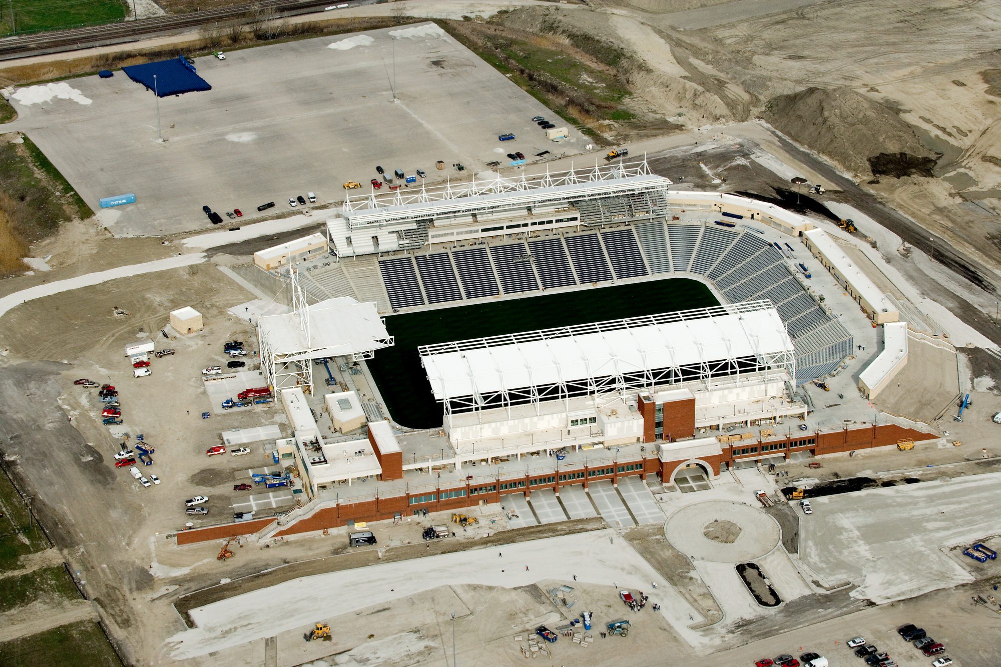 Aerial view of Bridgeview Stadium under construction, the seating bowl and brick concourse facade complete but the surrounding site still unpaved, with construction equipment across the grounds and no venue signage installed.